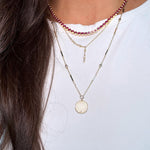 A close-up of a person wearing three layered necklaces: a gold St.Benedict coin pendant necklace, a dainty short asymmetrical lariat, and a pink tennis necklace necklace.