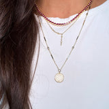 A close-up of a person wearing three layered necklaces: a gold St.Benedict coin pendant necklace, a dainty short asymmetrical lariat, and a pink tennis necklace necklace.